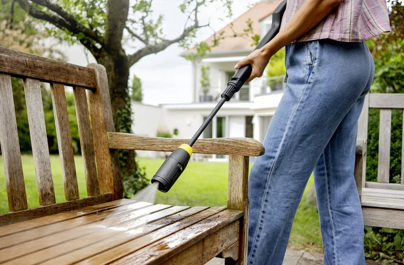 Person reinigt Holzbank im Garten mit Kärcher Hochdruckreiniger. Im Hintergrund ist ein modernes Haus zu sehen.