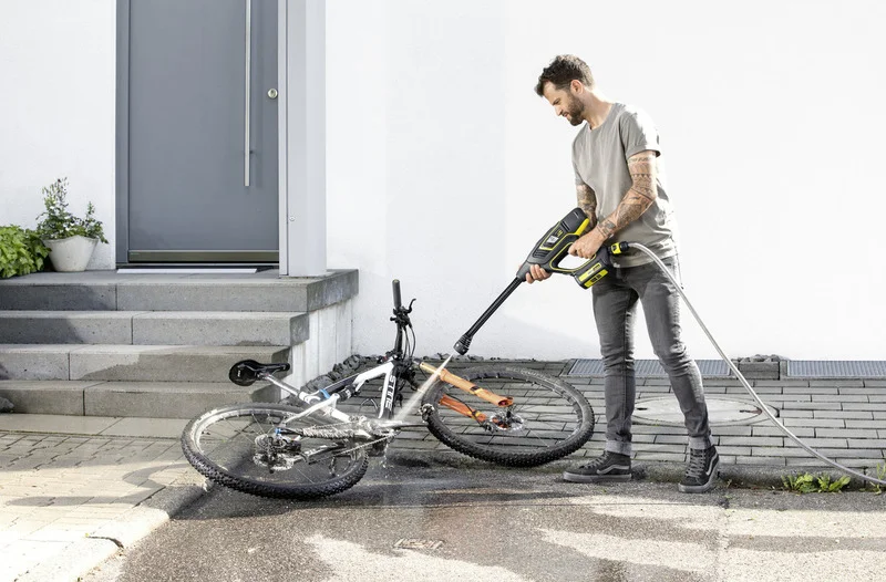 Mann reinigt Fahrrad mit Kärcher Hochdruckreiniger vor einer Haustür. Fahrrad liegt auf dem Boden, Umgebung ist gepflastert.