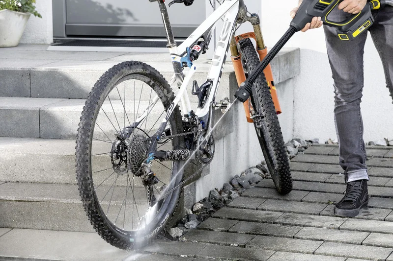 Person reinigt Fahrrad mit Kärcher Hochdruckreiniger auf gepflastertem Weg vor einer Treppe.