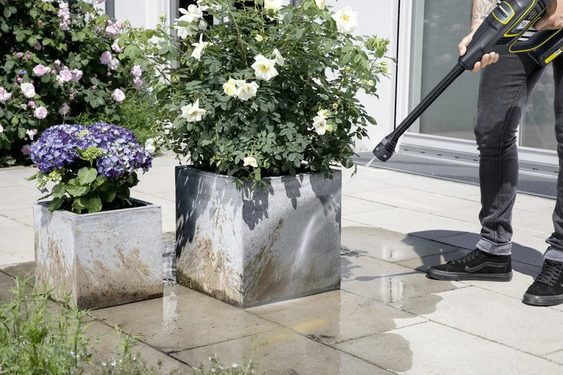 Person reinigt Pflanzkübel mit Kärcher Hochdruckreiniger auf Terrasse. Blumen im Hintergrund.