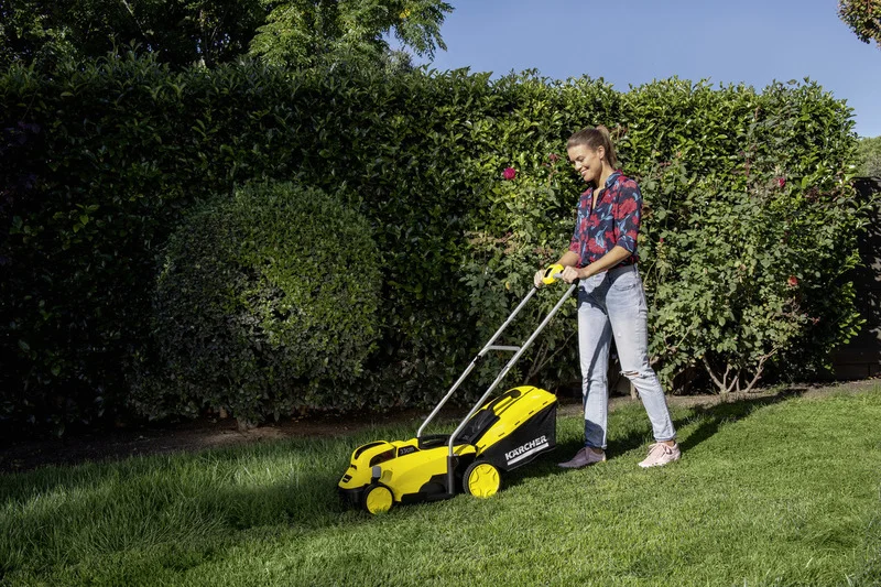 Person mäht Rasen mit einem Kärcher Rasenmäher im Garten, umgeben von grünen Hecken und Büschen.
