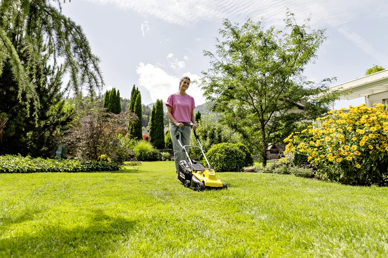 Person mäht Rasen mit einem gelben Kärcher Rasenmäher in einem blühenden Garten unter blauem Himmel.