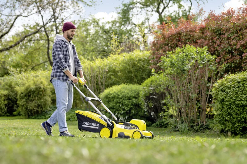 Person mäht Rasen mit einem Kärcher Rasenmäher in einem Garten, umgeben von Büschen und Bäumen.
