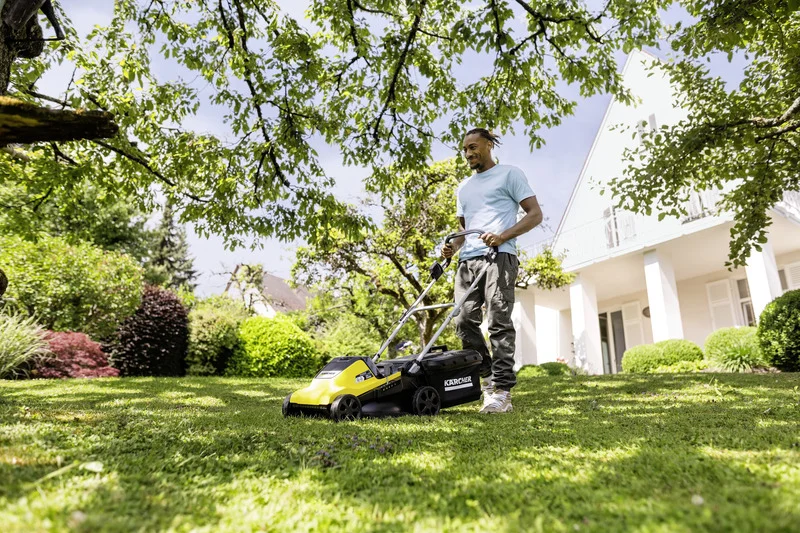 Person mäht Rasen mit einem Kärcher Rasenmäher im Garten, umgeben von Bäumen und einem modernen Haus im Hintergrund.