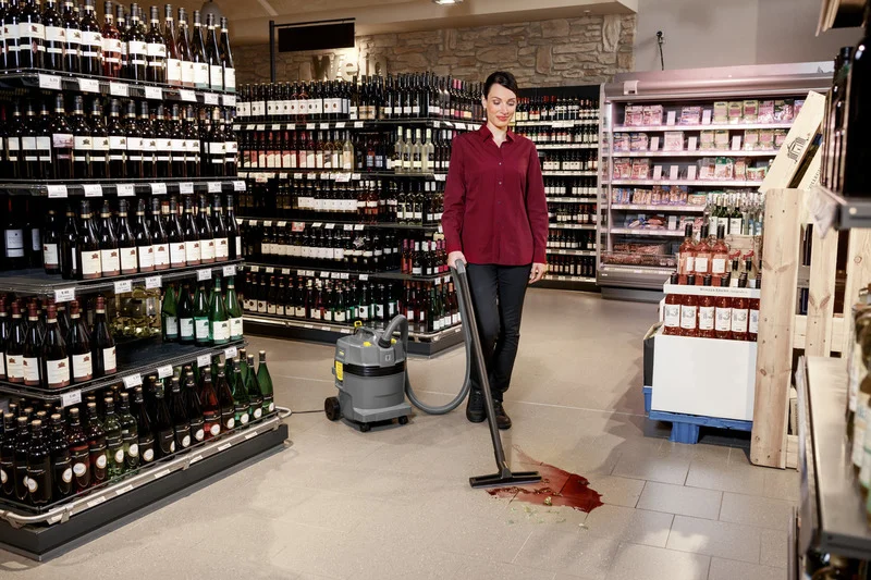 Person reinigt Rotweinfleck im Supermarkt mit Kärcher Nass-/Trockensauger. Umgebung voller Weinflaschen und Lebensmittelregale.