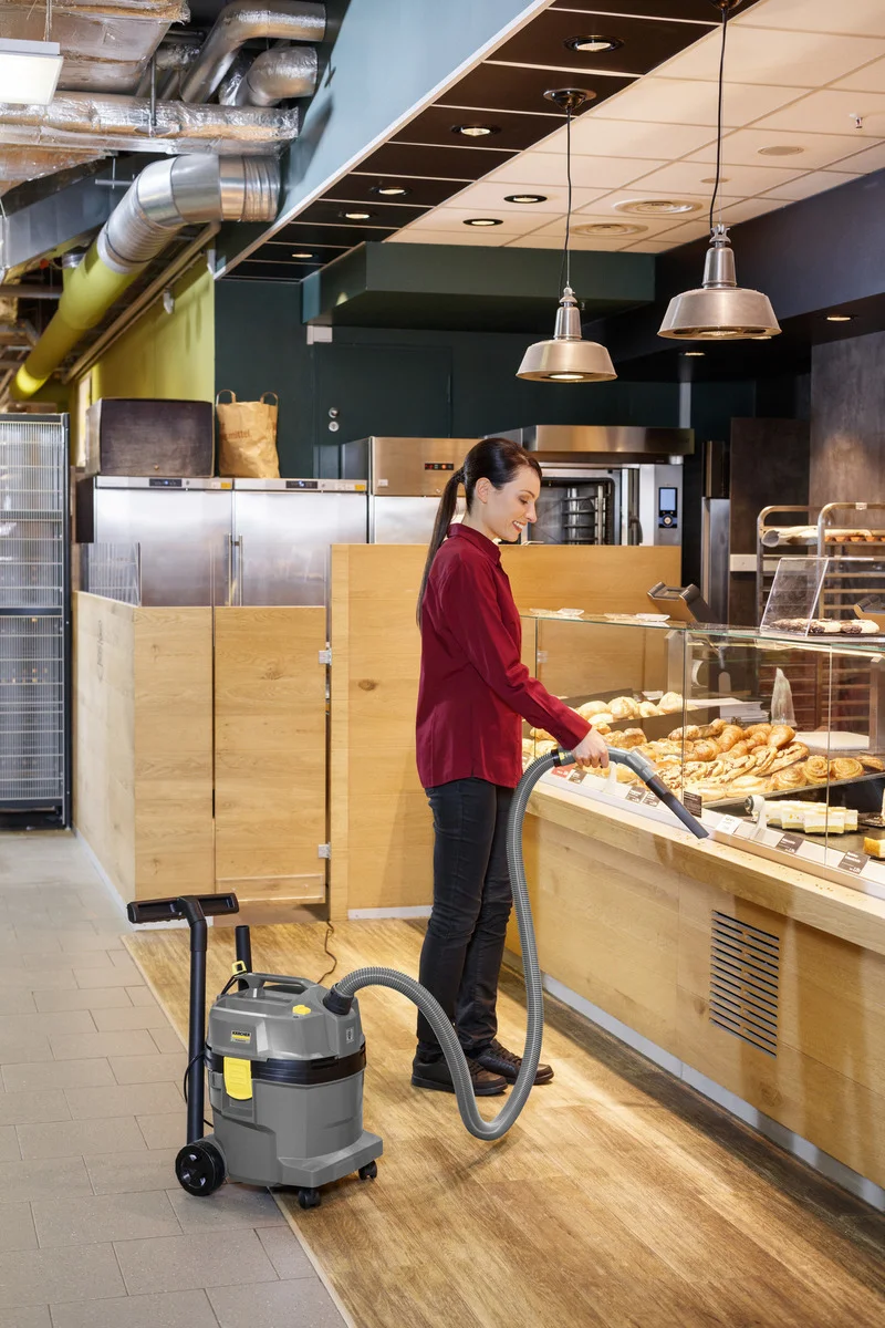 Frau in rotem Hemd reinigt Bäckereitheke mit Kärcher Nass-Trockensauger in modernem Laden. Holzverkleidung und Gebäck im Hintergrund.