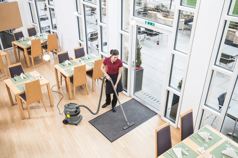Person reinigt Restaurantboden mit Kärcher Nass-Trockensauger. Holzboden, gedeckte Tische, große Fenster mit Blick nach draußen.