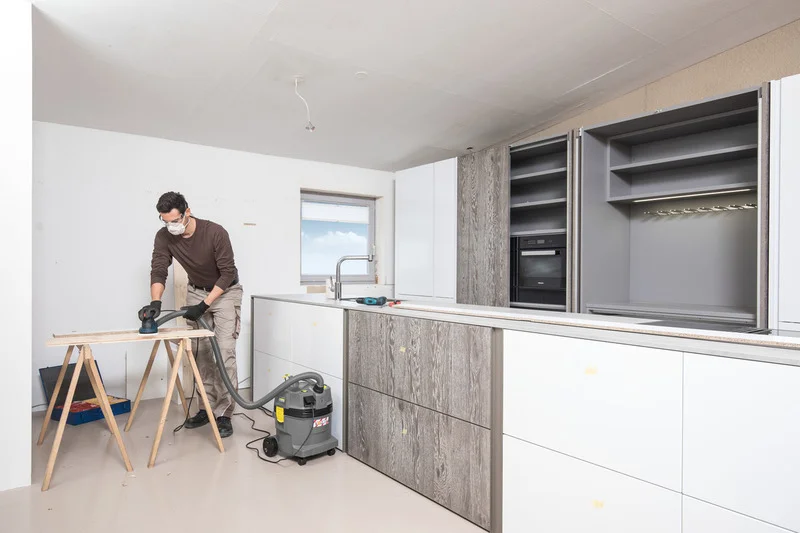 Mann schleift Holzplatte in moderner Küche. Kärcher Nass-/Trockensauger steht daneben. Schränke und Fenster im Hintergrund.