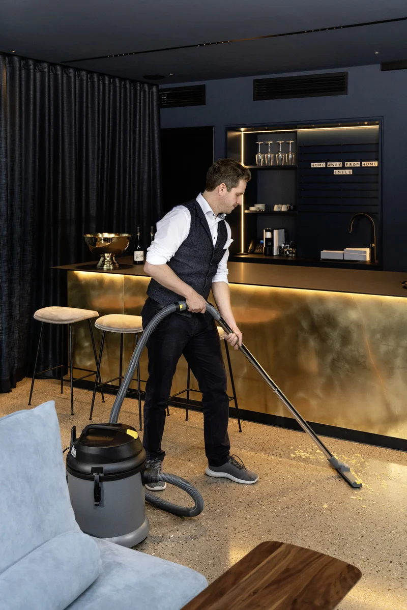 Mann in eleganter Kleidung saugt mit einem Kärcher Nass-/Trockensauger den Boden in einer stilvollen Bar mit goldener Theke.