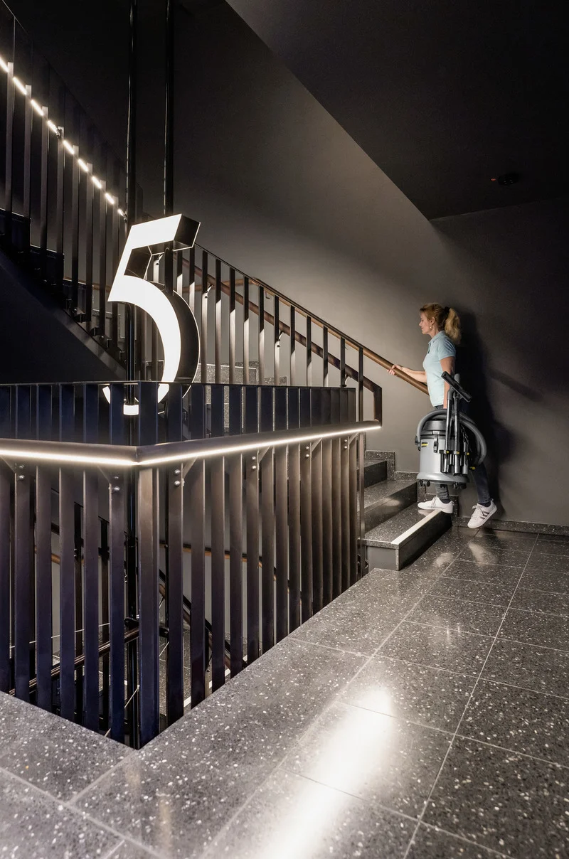 Person trägt Kärcher Nass-/Trockensauger eine Treppe hinauf in einem modernen Gebäude mit beleuchteter Zahl 5.