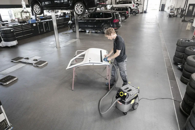 Mann bearbeitet Autoteil in Werkstatt mit Kärcher Nass-Trockensauger. Autos auf Hebebühnen, Reifenstapel im Hintergrund.
