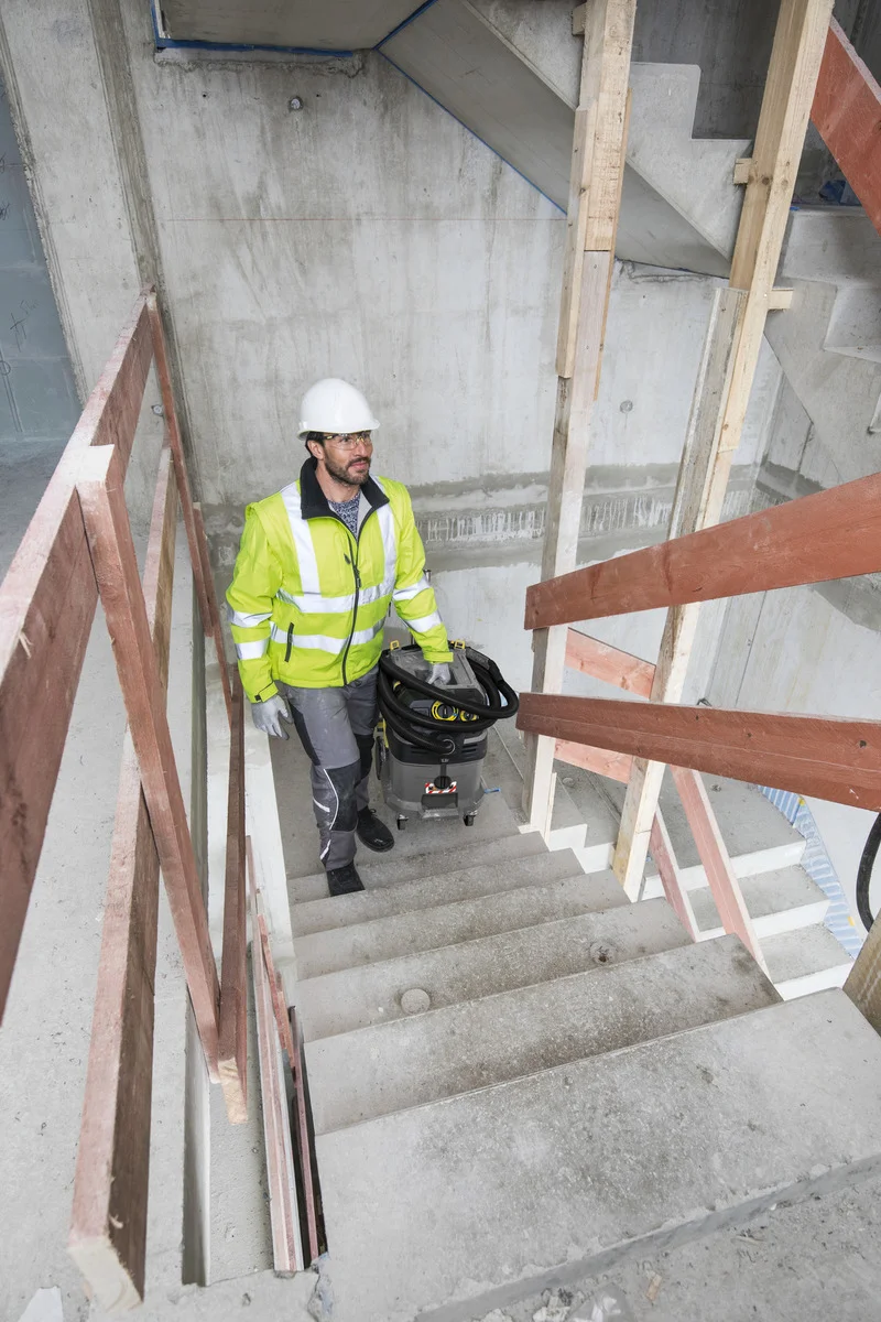 Person in Sicherheitskleidung und Helm trägt einen Kärcher Industriesauger eine Betontreppe in einem Rohbau hinauf.