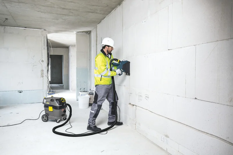 Arbeiter in Schutzkleidung nutzt Wandfräse in Rohbau. Kärcher Industriesauger steht daneben. Raum ist hell und unfertig.