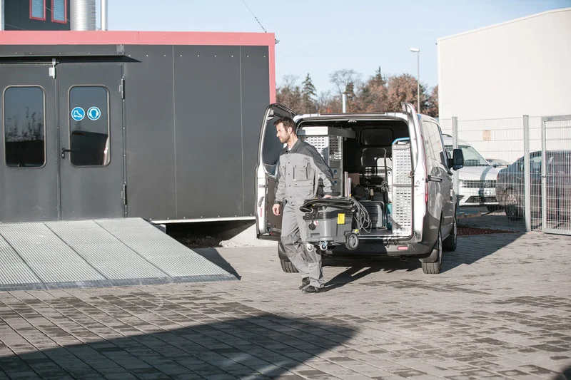 Mann in Arbeitskleidung transportiert Kärcher Nass-/Trockensauger aus offenem Lieferwagen auf gepflastertem Hof. Gebäude im Hintergrund.