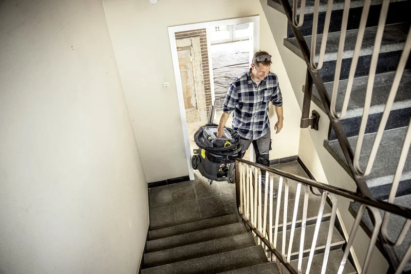 Mann trägt Kärcher Nass-/Trockensauger die Treppe hinauf in einem renovierten Gebäude mit offener Tür und Baustellenmaterialien.