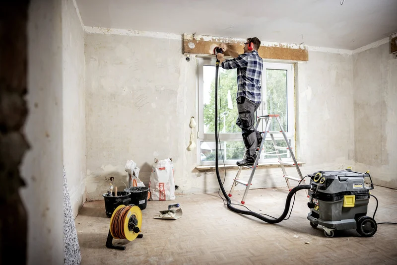 Handwerker auf Leiter bei Renovierung, verwendet Elektrowerkzeug. Kärcher Nass-/Trockensauger und Kabeltrommel im Raum. Wand und Fenster sichtbar.