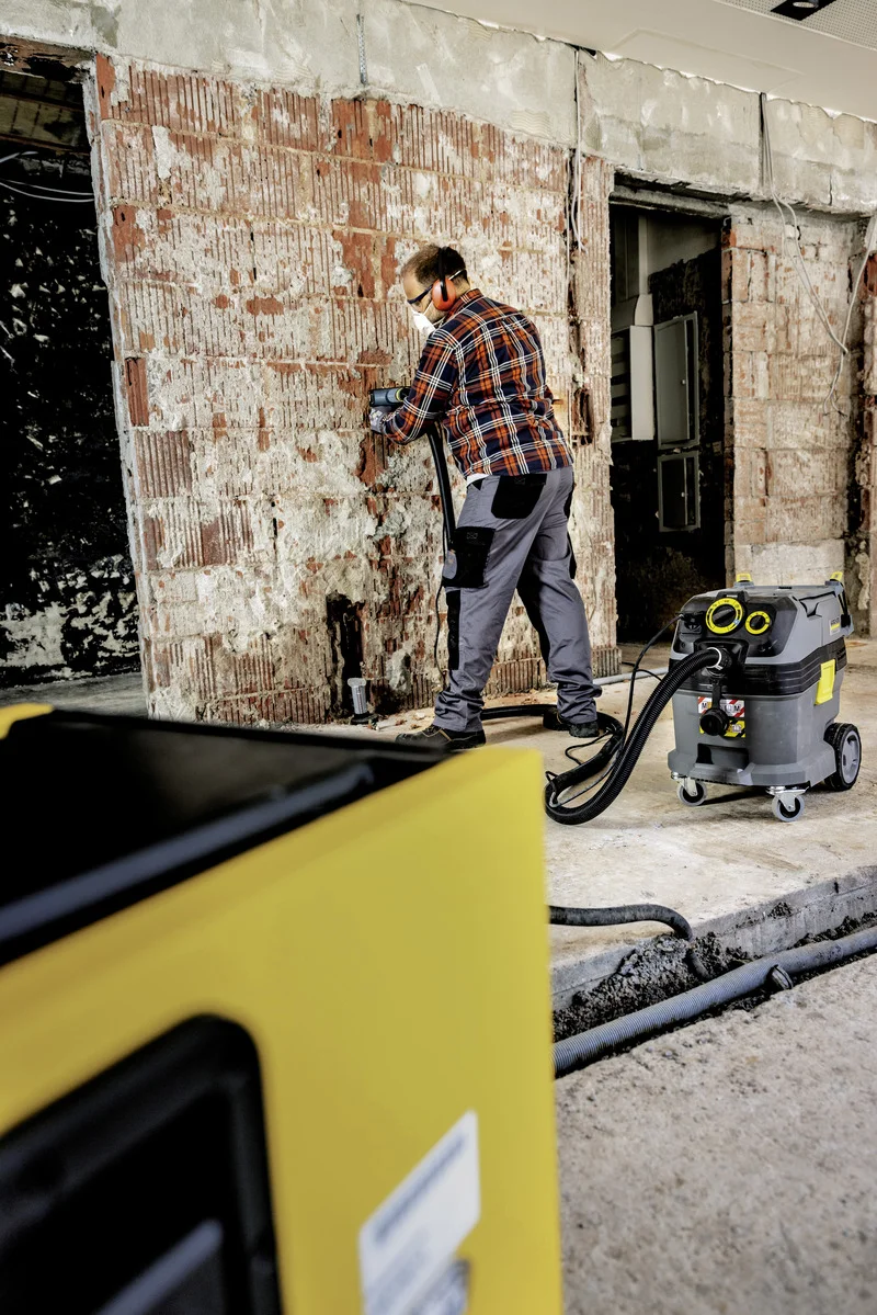 Person arbeitet an einer Wand in einem Rohbau. Ein Kärcher Industriesauger steht daneben. Im Vordergrund ein gelbes Gerät.