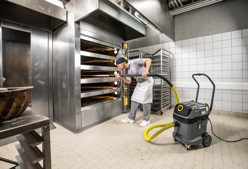 Bäcker reinigt Backofen mit Kärcher Nass-/Trockensauger in einer gewerblichen Küche. Edelstahlöfen und Backblechregale im Hintergrund.