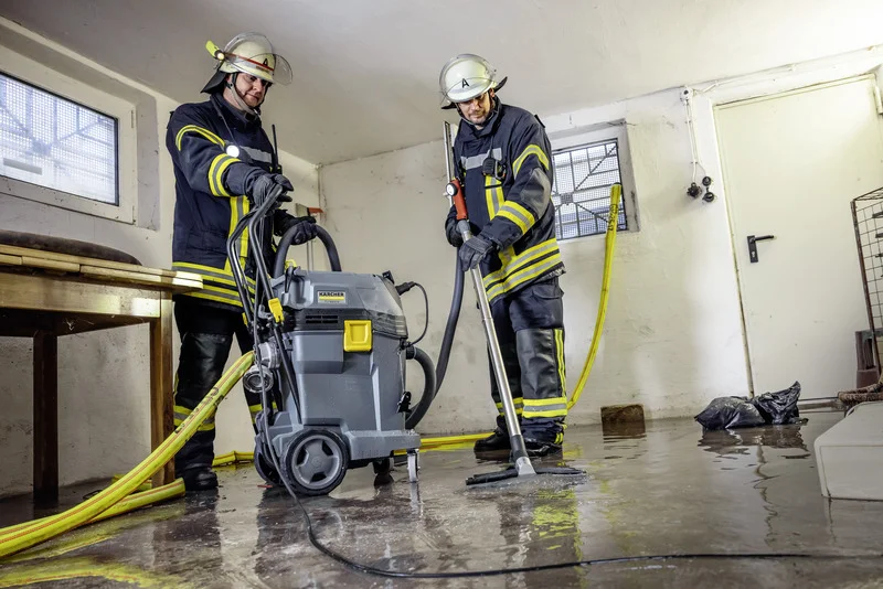 Zwei Feuerwehrleute in Schutzkleidung nutzen einen Kärcher Nass-/Trockensauger in einem überschwemmten Raum.
