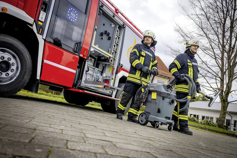 Zwei Feuerwehrleute in Schutzkleidung stehen neben einem Feuerwehrfahrzeug und einem Kärcher Nass-/Trockensauger auf einem gepflasterten Weg.