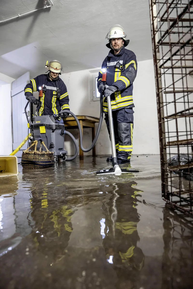 Feuerwehrleute in Schutzkleidung beseitigen Wasser in einem Keller mit einem Kärcher Nasssauger. Umgebung ist überschwemmt.