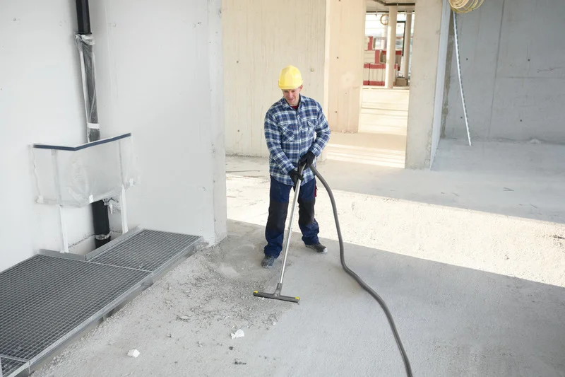 Bauarbeiter mit gelbem Helm reinigt Betonboden mit einem Kärcher Industriesauger in einem unfertigen Gebäude.