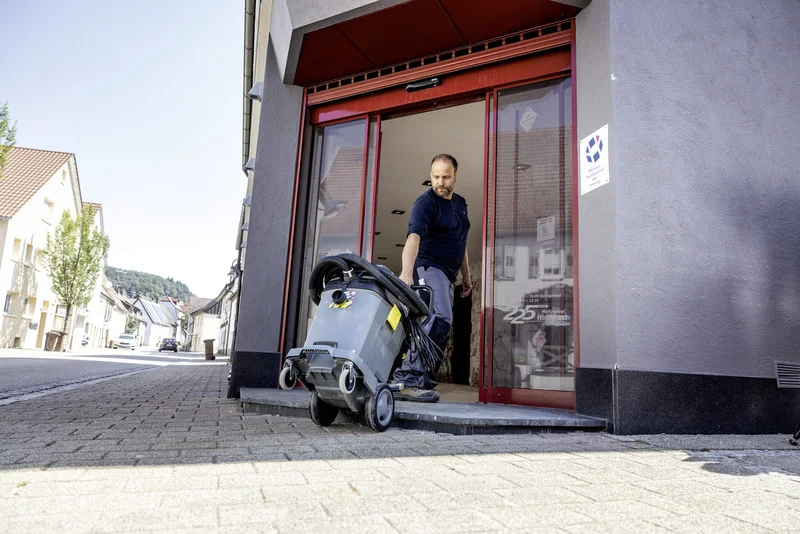 Person schiebt Kärcher Nass-/Trockensauger aus einem Gebäude mit rotem Türrahmen auf eine gepflasterte Straße in einer städtischen Umgebung.