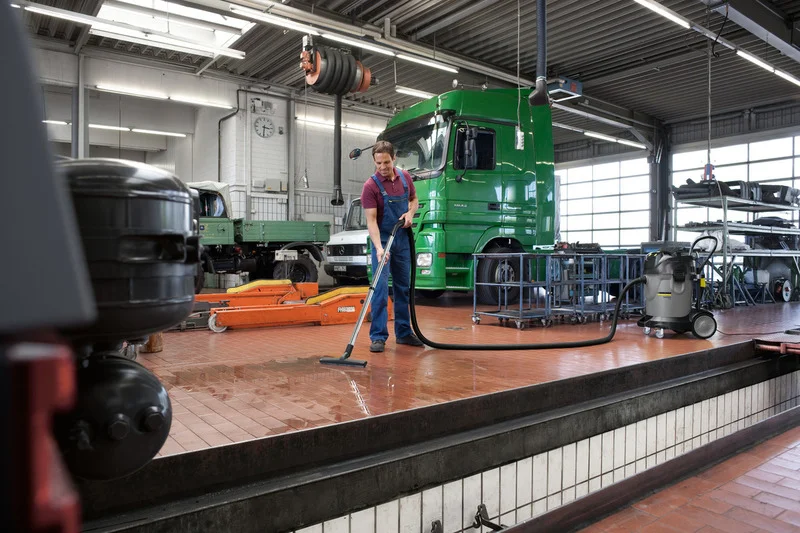 Mann reinigt Boden in Werkstatt mit Kärcher Nass-/Trockensauger. Im Hintergrund grüner LKW und Hebebühne. Industrielle Umgebung.