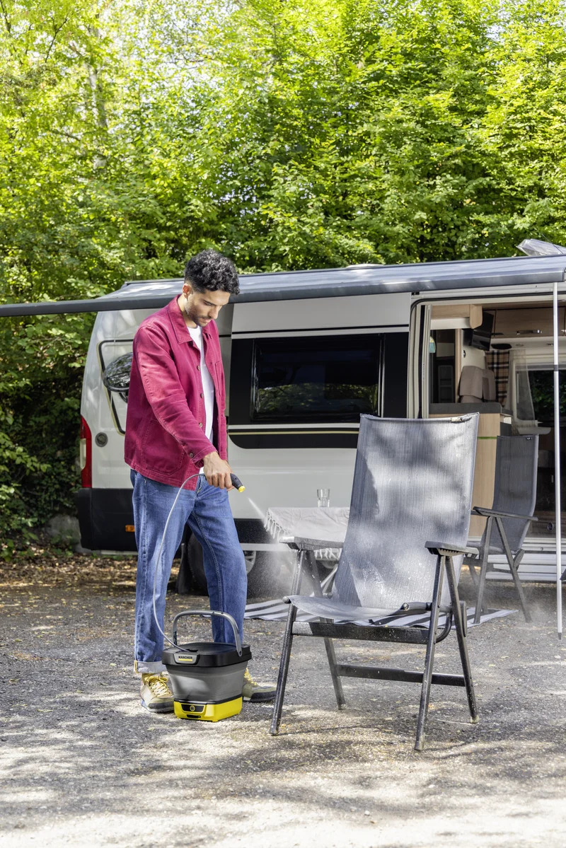 Person reinigt Campingstuhl mit Kärcher Hochdruckreiniger, Wohnmobil im Hintergrund, sonniger Tag, grüne Bäume umgeben die Szene.