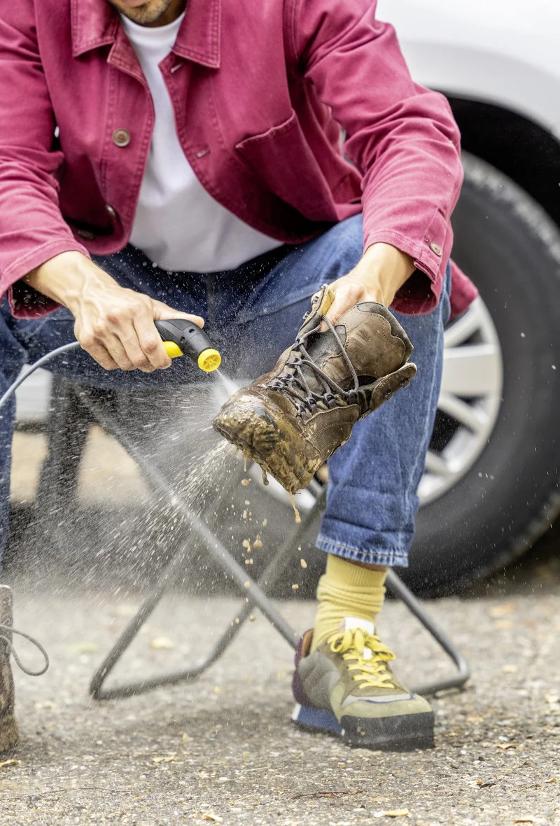 Person reinigt schmutzigen Wanderschuh mit Kärcher Hochdruckreiniger im Freien. Sitzend auf einem Klappstuhl, Auto im Hintergrund.