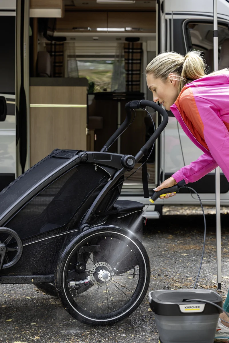 Frau reinigt Kinderwagenrad mit Kärcher Hochdruckreiniger, im Hintergrund Wohnmobil, Eimer mit Kärcher-Logo auf dem Boden.