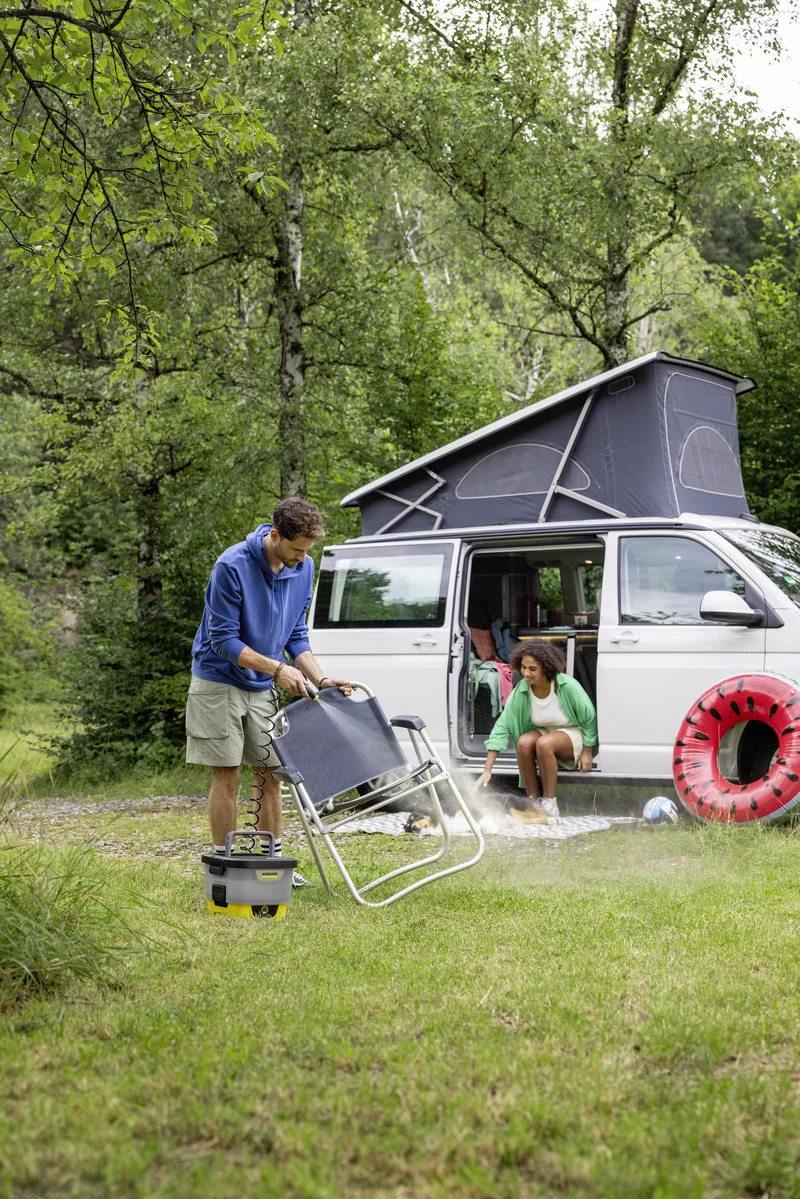 Person reinigt Campingstuhl mit Kärcher Niederdruckreiniger neben Camper-Van im Wald. Zweite Person sitzt im Van, Wassermelonen-Schwimmring daneben.