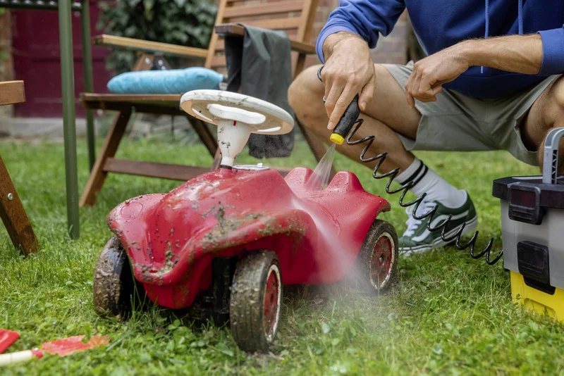Person reinigt rotes Kinderauto im Garten mit Kärcher Hochdruckreiniger. Im Hintergrund Gartenmöbel und Werkzeugkasten.