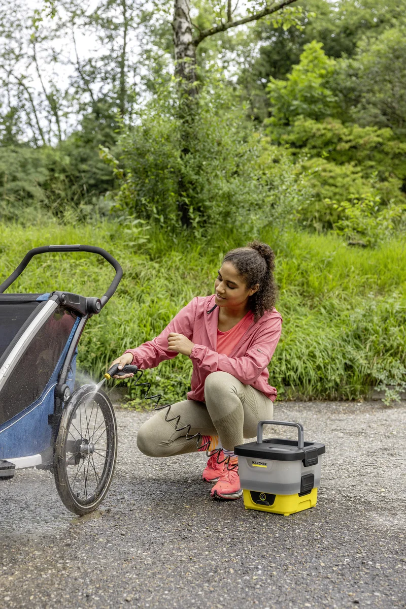 Person reinigt Kinderwagenrad mit Kärcher Niederdruckreiniger, draußen auf einem Weg, umgeben von grüner Vegetation.