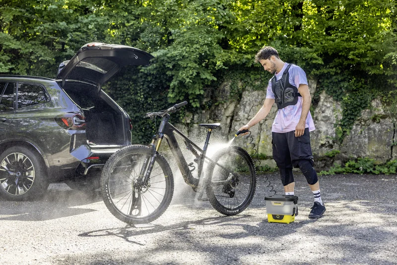 Person reinigt Fahrrad mit Kärcher Hochdruckreiniger neben einem Auto im Freien, umgeben von Bäumen und Felsen. Kofferraum des Autos ist offen.