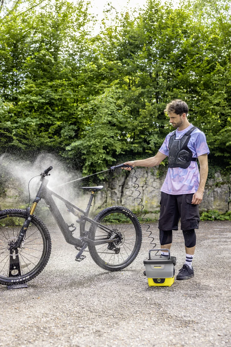 Mann reinigt ein Mountainbike mit einem Kärcher Hochdruckreiniger im Freien, umgeben von grünen Bäumen und einer Steinmauer.