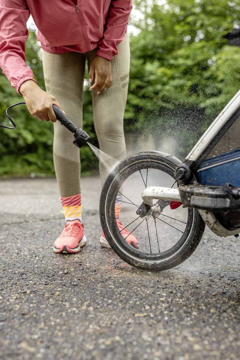 Person reinigt Fahrradreifen mit Kärcher Hochdruckreiniger im Freien. Trägt rote Jacke, beige Leggings und bunte Socken.
