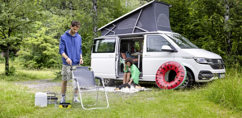 Mann sprüht Wasser auf Campingstuhl mit Kärcher Gerät, neben ihm ein Hund und ein Kind vor einem weißen Camper im Wald.