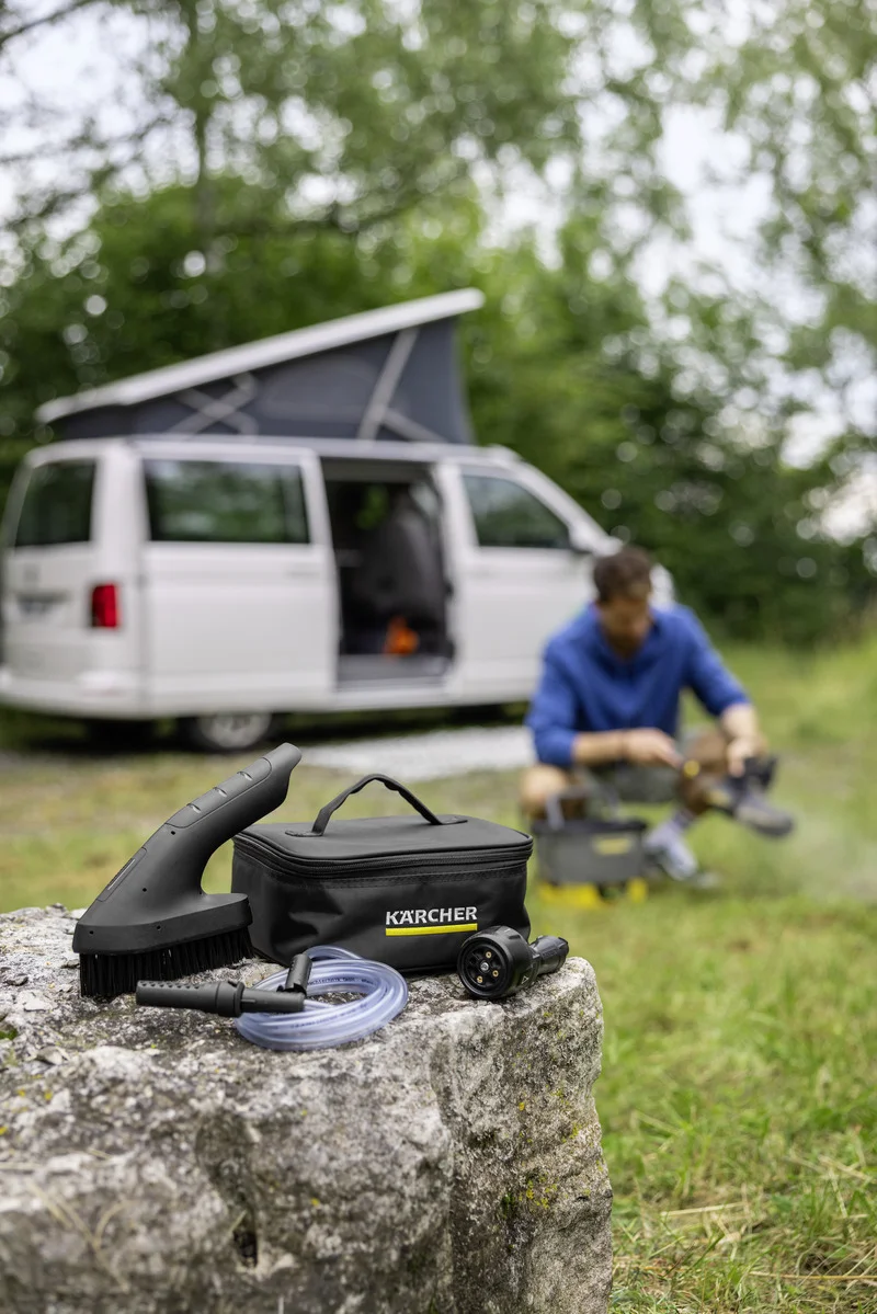 Kärcher Reinigungsset auf einem Stein im Freien, im Hintergrund ein geöffneter Campingbus und eine Person beim Werkeln.