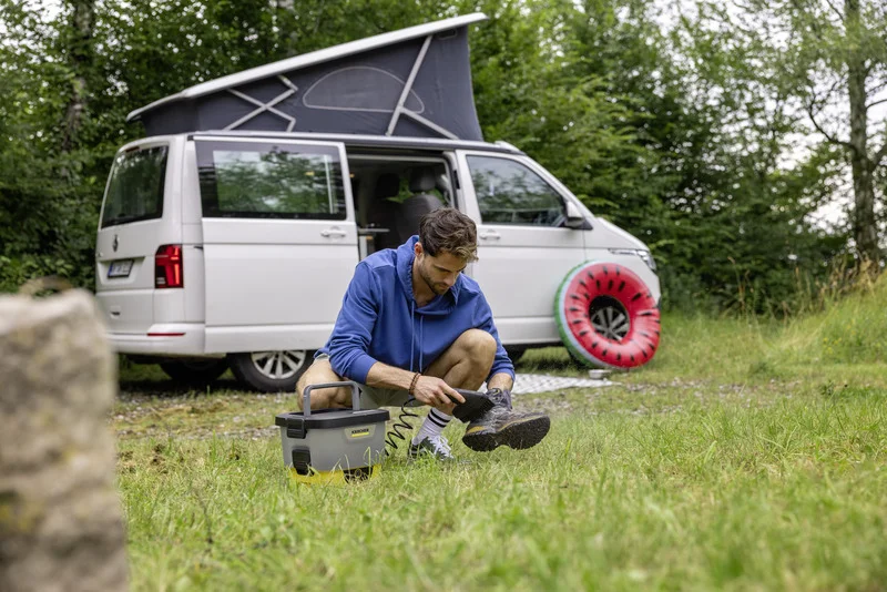 Mann reinigt Schuhe mit Kärcher Niederdruckreiniger vor einem weißen Camper im Freien. Ein Wassermelonen-Schwimmring liegt daneben.