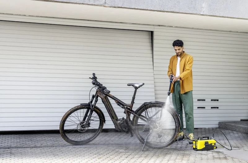 Person reinigt ein schmutziges Fahrrad mit einem Kärcher Hochdruckreiniger vor einer geschlossenen Garagentür.