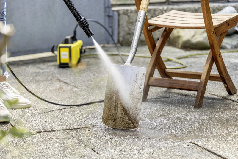 Kärcher Hochdruckreiniger reinigt eine schmutzige Schaufel auf einer Terrasse neben einem Holzstuhl.