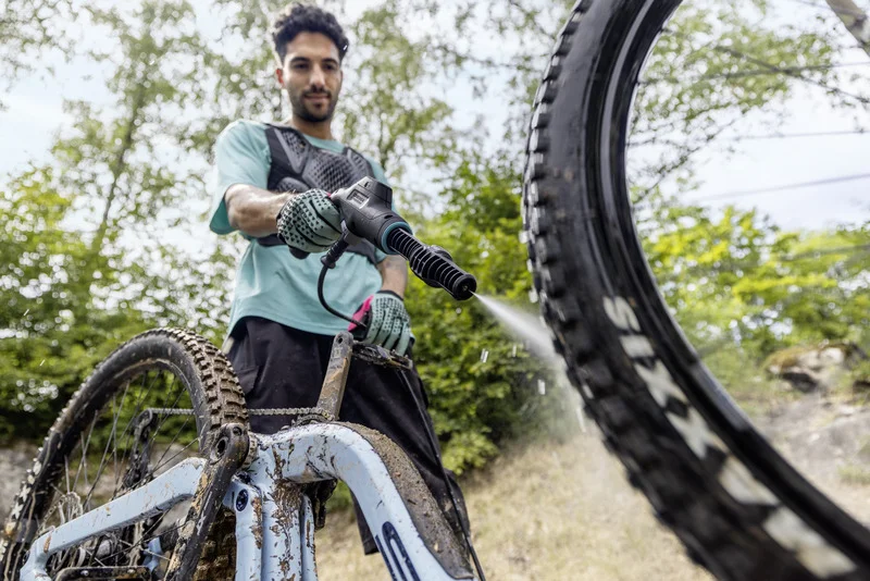 Person reinigt ein schmutziges Fahrrad mit einem Kärcher Hochdruckreiniger im Freien, umgeben von Bäumen und Gras.