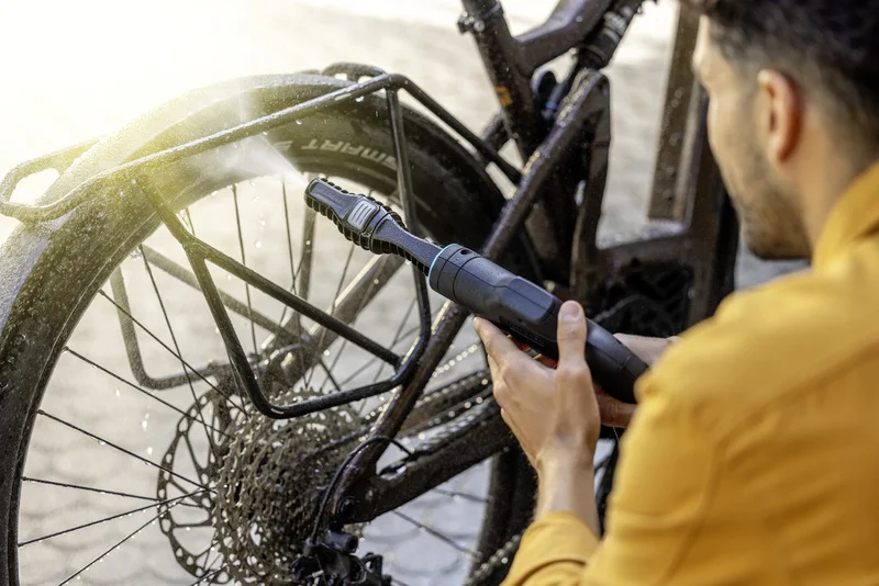 Person reinigt Fahrrad mit Kärcher Hochdruckreiniger, Fokus auf Hinterrad und Kette, sonnige Umgebung.