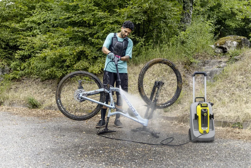 Person reinigt ein schmutziges Fahrrad mit einem Kärcher Hochdruckreiniger im Freien, umgeben von Bäumen und Gras.