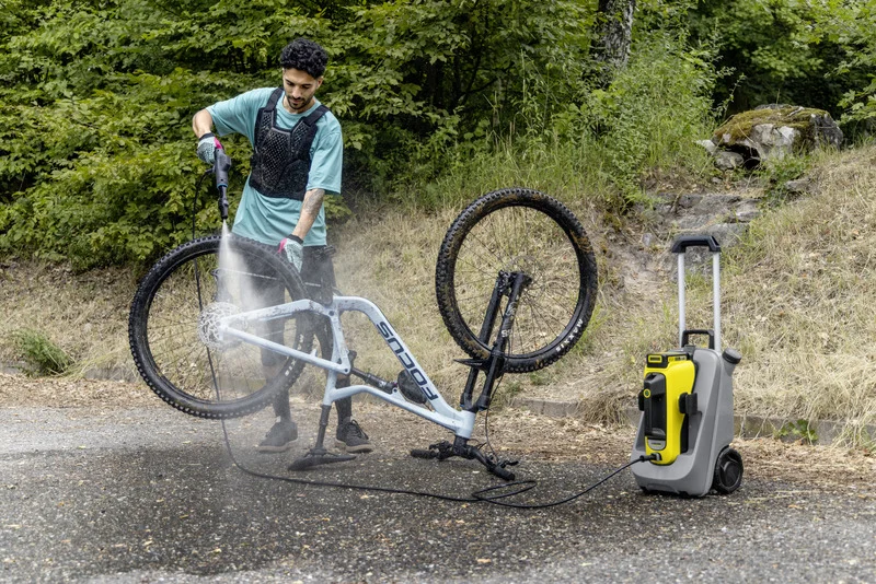 Person reinigt Fahrrad mit Kärcher Hochdruckreiniger im Freien. Fahrrad steht auf Kopfsteinpflaster, umgeben von Bäumen und Gras.