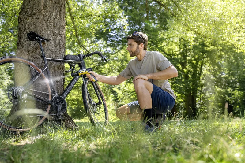 Mann reinigt Fahrrad mit Kärcher Hochdruckreiniger im Freien neben einem Baum, umgeben von grünem Gras und Bäumen.