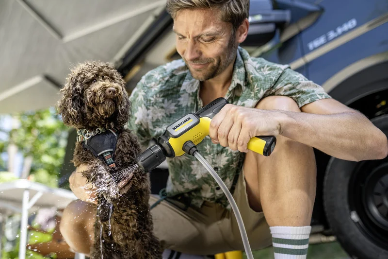 Mann reinigt Hund mit Kärcher Reinigungsgerät im Freien, neben einem Fahrzeug. Hund trägt ein Geschirr, Wasser spritzt aus dem Gerät.