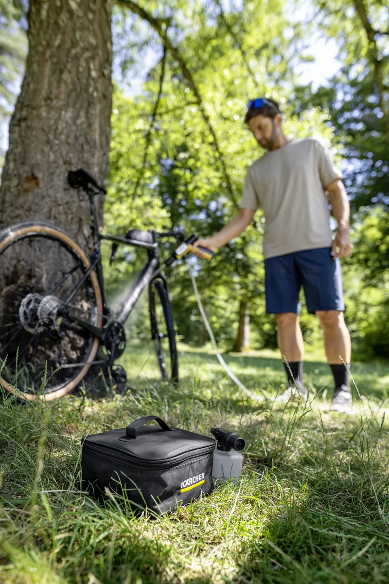 Mann reinigt Fahrrad mit Kärcher Hochdruckreiniger im Freien. Fahrrad lehnt an Baum, Kärcher Tasche im Gras.