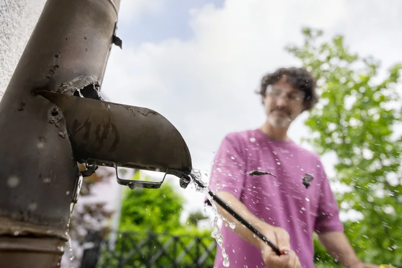 Person in rosa T-Shirt reinigt ein verrostetes Rohr im Freien mit einem Hochdruckreiniger. Wasser spritzt aus dem Rohr.